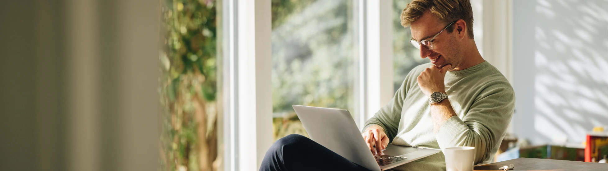  Young man in glasses and a light sweater working happily on a laptop at a wooden table in a bright home office, with large windows overlooking greenery.